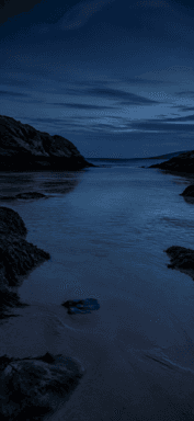 A calm blue cove lies between dark rocks beneath a cloudy night sky.