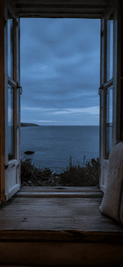 An open window above a cushioned ledge looks out towards the sea and a cloudy blue sky.
